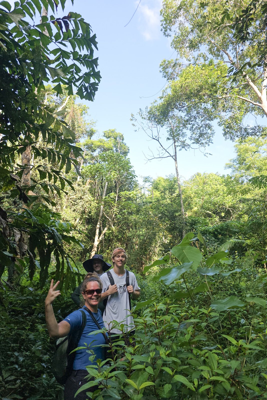 Trio of trekkers hiking through dense jungle, one giving peace sign