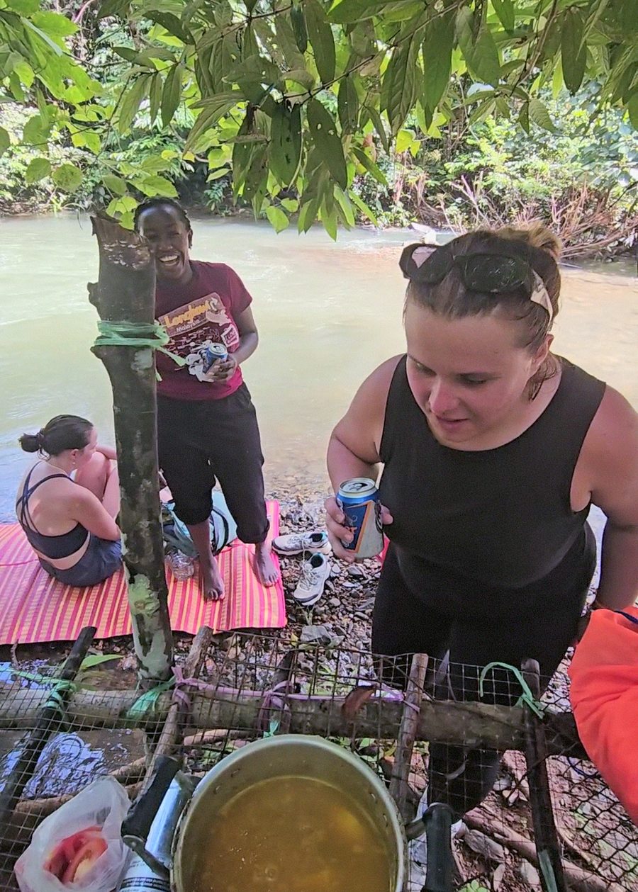 Friends gathered at the handmade Jungle Bar table with cold drinks and a fresh pot of curry by the water