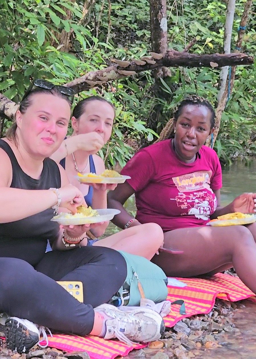 Three friends eating freshly cooked lunch — rice and curry — by the water after the trek