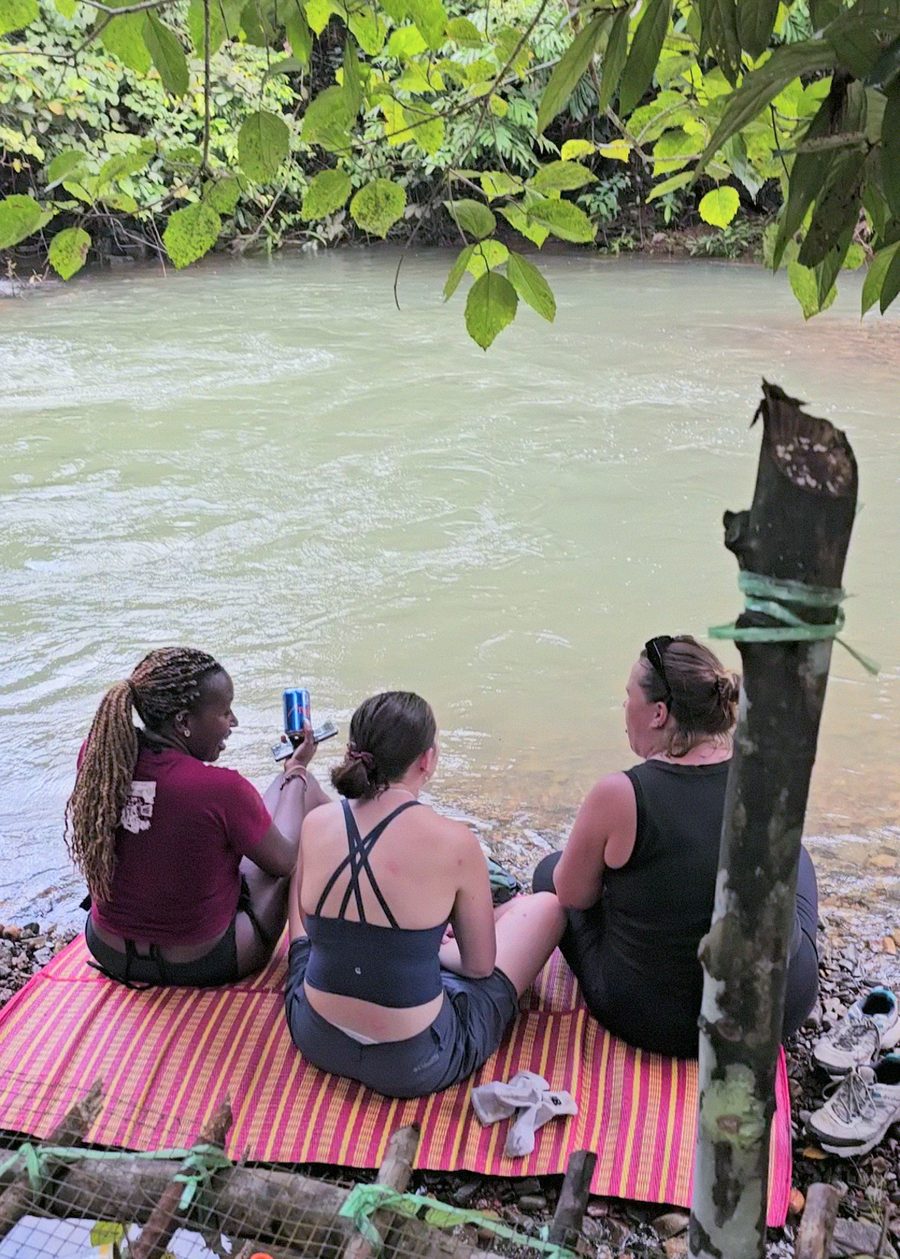 Three friends sitting on a woven mat by the water, sharing drinks and stories